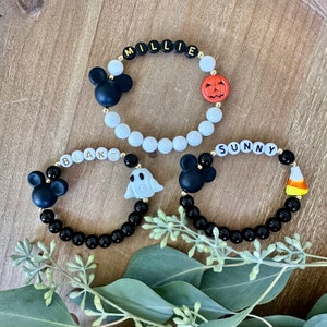 Puede incluir: Tres pulseras de cuentas con nombres personalizados y dijes temáticos de Halloween. Una pulsera tiene un dije de cabeza de Mickey Mouse y el nombre "MILLIE". Otra tiene un dije de fantasma y el nombre "BLAKE". La tercera tiene un dije de cabeza de Mickey Mouse y el nombre "SUNNY".