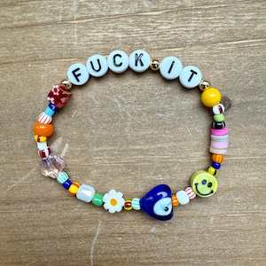 May include: A beaded bracelet with the word "FUCK IT" spelled out in white letter beads. The bracelet features a variety of colorful beads, including a blue heart-shaped bead, a smiley face bead, and flower beads. The bracelet is on a wooden surface.