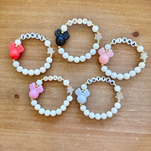 May include: Five white beaded bracelets with gold lettering spelling out names, each with a different colored Mickey Mouse charm. The names are Jones, Karis, Fiona, Beau, and Sutter.