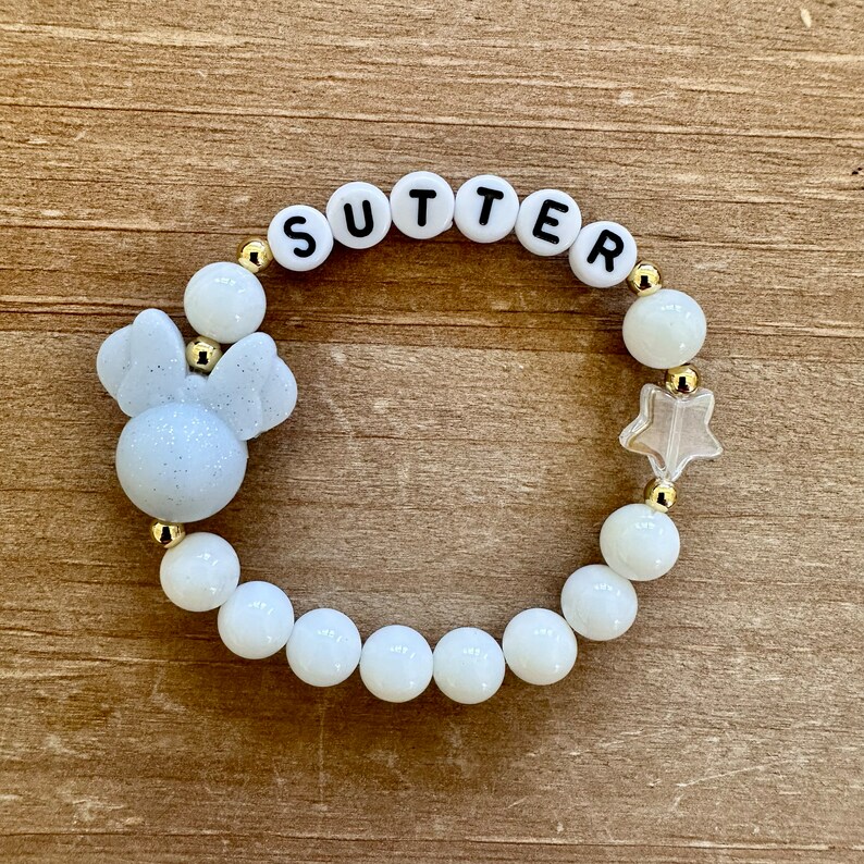 May include: White beaded bracelet with gold accents, featuring the name "SUTTER" spelled out in black letters, a white sparkly Mickey Mouse head charm, and a clear star charm.