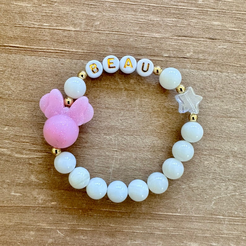 May include: A white beaded bracelet with gold accents, featuring the name "BEAU" spelled out in white beads, a pink glitter Mickey Mouse charm, and a clear star charm.
