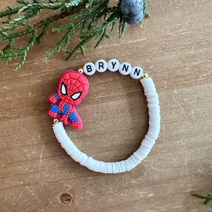 May include: White beaded bracelet with a gold charm and a red and blue Spiderman character charm. The bracelet has the name "BRYNN" spelled out in black letters on white beads.