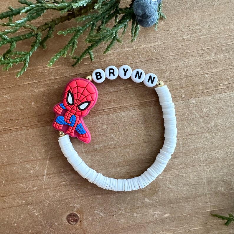 May include: White beaded bracelet with a gold charm and a red and blue Spiderman character charm. The bracelet has the name "BRYNN" spelled out in black letters on white beads.