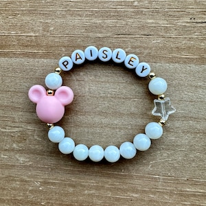 May include: A white and gold beaded bracelet with the name "Paisley" spelled out in white letters. The bracelet also features a pink Mickey Mouse charm and a clear star charm.