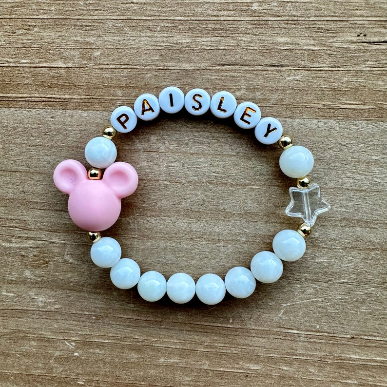 May include: A white and gold beaded bracelet with the name "Paisley" spelled out in white letters. The bracelet also features a pink Mickey Mouse charm and a clear star charm.