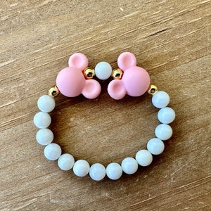 May include: A pink silicone bracelet with two mouse ears and gold accents. The bracelet is made with white beads.