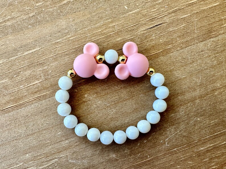May include: A pink silicone bracelet with two mouse ears and gold accents. The bracelet is made with white beads.