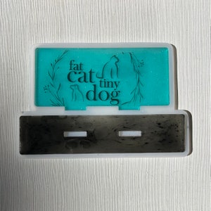 May include: A rectangular, two-part container with a teal interior and a black base. The teal section features the words "fat cat tiny dog" with cat and dog silhouettes. The container is white-rimmed and sits on a white surface.
