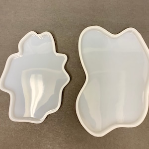 May include: Two clear, irregularly shaped silicone molds with white borders. The molds have a glossy finish and are designed for crafting projects. The shapes are organic and abstract, suitable for resin art or other DIY creations.
