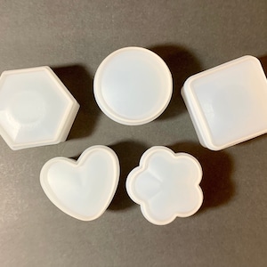 May include: Five white silicone molds in different shapes: a hexagon, a circle, a square, a heart, and a flower. The molds are designed for making resin crafts.