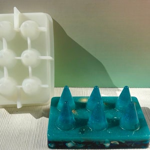May include: A silicone mold for making resin crafts. The mold is white and has six cone-shaped cavities. A finished resin piece is shown in the foreground. The resin piece is teal blue and has a glittery finish. It has six cone-shaped protrusions.