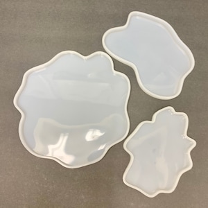 May include: Three white silicone molds in different irregular shapes. The molds are designed for resin casting and are perfect for creating unique coasters or decorative trays.