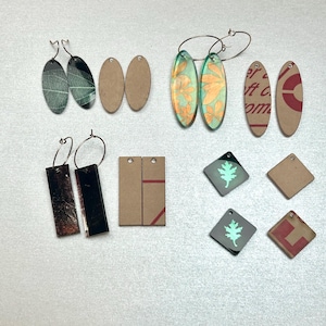May include: A collection of handmade earrings in various shapes and designs. Includes oval, rectangular, and diamond-shaped earrings. Some earrings feature leaf patterns, floral designs, and geometric shapes. Materials include metal, paper, and glow-in-the-dark elements.