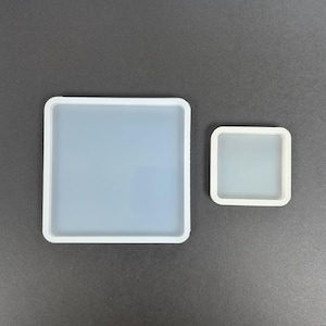 May include: Two square silicone molds with rounded corners, featuring a translucent, semi-opaque center and a solid white border. The larger mold is positioned on the left, while the smaller mold is placed to its right, both displaying a smooth finish.