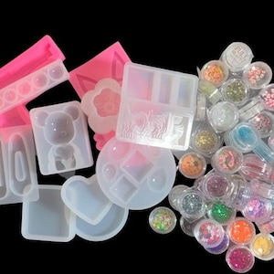 May include: Assortment of clear plastic containers filled with colorful glitter and sequins.  The containers are arranged in a pile on a black background.  There are also several pink and clear silicone molds in the image.