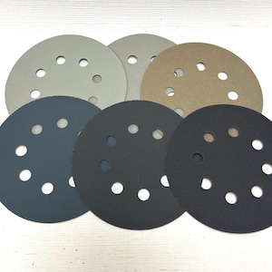 May include: Seven circular sanding discs in shades of grey, tan, and black. Each disc features multiple evenly spaced holes. The discs are arranged in a slightly overlapping pattern against a white background.