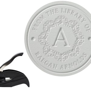 May include: A black self-inking stamp with a silver handle. The stamp design is a circle with the text "From the Library of Raegan Arnolds" and a decorative flourish with the letter "A" in the center.