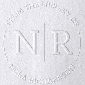 May include: A white paper surface featuring a circular blind embossed design. The text reads FROM THE LIBRARY OF at the top and NORA RICHARDSON at the bottom, with the letters N and R separated by a vertical line in the center of the circle.