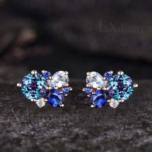 May include: A pair of rose gold stud earrings with a cluster of blue and white gemstones. The earrings feature a mix of round and oval-shaped stones, including blue topaz, aquamarine, and alexandrite.