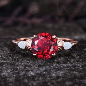May include: A rose gold engagement ring with a large, round, red gemstone surrounded by two smaller, pear-shaped, white gemstones.