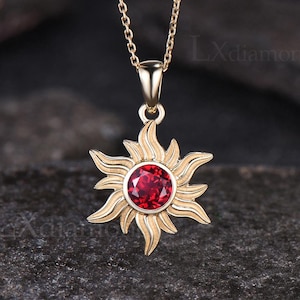 May include: A gold necklace with a red gemstone sun pendant. The pendant features a detailed sun design with a round red gemstone in the centre.