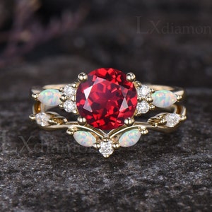 Puede incluir: Un anillo de oro con una gran piedra roja rodeada de piedras blancas más pequeñas. La banda está adornada con ópalos blancos y más piedras blancas pequeñas. El anillo está sobre un fondo gris.