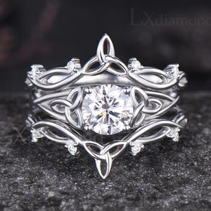 May include: A three-piece silver ring set with a Celtic knot design. The engagement ring features a large, round, clear gemstone. The bands are adorned with smaller accent stones and intricate knotwork. The rings are displayed on a dark, textured surface.