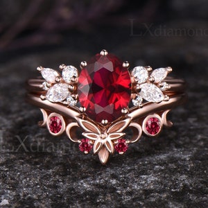 Puede incluir: Un anillo de compromiso de oro rosa con una gran piedra de rubí roja rodeada de pequeños diamantes blancos. El anillo presenta un delicado diseño floral con un motivo de mariposa.