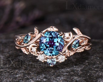 Puede incluir: Un anillo de oro rosa con una gran piedra preciosa redonda azul y morada rodeada de pequeñas piedras preciosas redondas blancas y azules. El anillo presenta un delicado diseño floral con hojas y enredaderas.