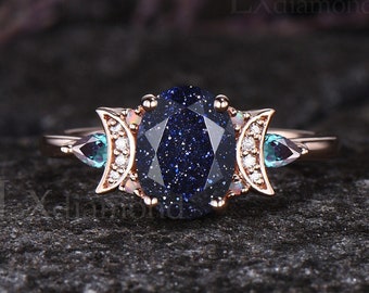 Peut inclure: Une bague en or rose avec une grande pierre précieuse ovale bleue entourée de petites pierres blanches. La bague présente deux motifs de croissant de lune de chaque côté de la pierre principale, chacun avec une petite pierre bleue.