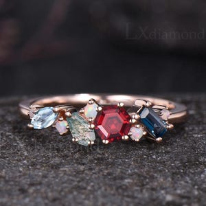 May include: A rose gold ring featuring a cluster of colorful gemstones. The ring showcases a central red hexagonal stone, surrounded by blue, green, and white accent stones. The ring is set on a dark surface.