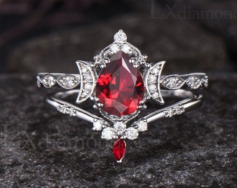 May include: A pear-shaped red gemstone ring with a delicate, intricate design featuring two crescent moons and a row of smaller gemstones. The ring is set in silver and has a matching band with a V-shaped design.