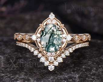 May include: A gold-toned engagement ring set with a large, oval-cut moss agate stone, surrounded by small diamonds. The band features intricate detailing and a matching diamond-studded wedding band. The ring set is displayed on a dark, textured surface.