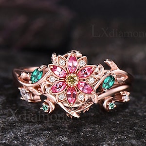 May include: A rose gold ring featuring a central flower motif with pink marquise-cut stones, a yellow center stone, and small clear accent stones. The band is designed with vine-like details and includes small green marquise-cut stones.