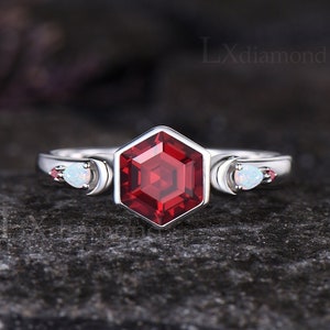 May include: A silver ring with a red hexagonal gemstone and two smaller white gemstones on either side. The ring is set on a dark gray background.