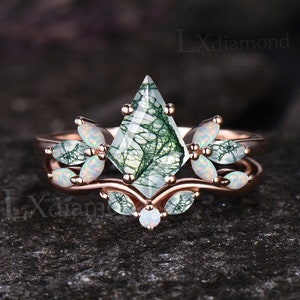 May include: A rose gold ring set with a moss agate gemstone and opal accents. The moss agate is a kite-shaped stone with a green and white pattern. The opal accents are set in a floral design.