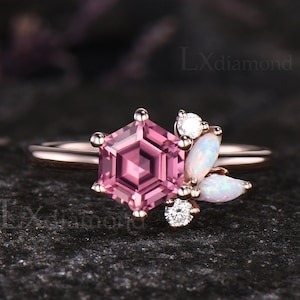 May include: A rose gold ring featuring a central hexagonal pink gemstone held by six prongs. To the side, a cluster of two round clear stones and two marquise-shaped opals are set. The band is smooth and polished. The text LXdiamond is visible.