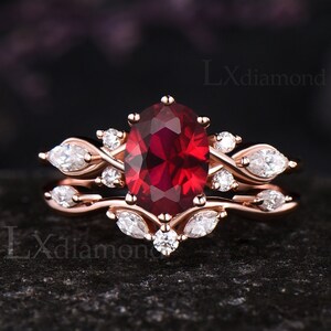 May include: Rose gold engagement ring set with a large oval-cut, deep red gemstone and smaller white accent stones. The band features a delicate, intertwined design. The ring is set against a dark background.