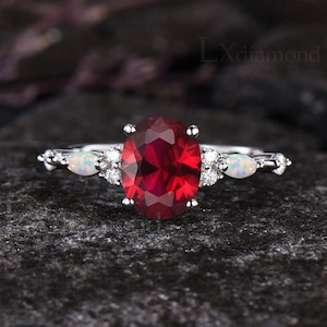 May include: A silver ring with a large oval red gemstone surrounded by smaller white gemstones. Two smaller white gemstones are set on either side of the main stone.