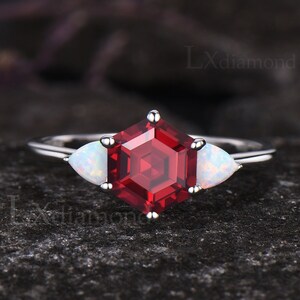 May include: A silver ring featuring a hexagonal, faceted, deep red gemstone as the centerpiece, flanked by two pear-shaped, white opal stones. The ring is set against a dark, textured background.