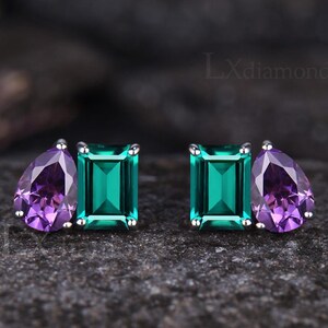 May include: Stud earrings with a pear-shaped purple gemstone and an emerald-cut green gemstone. The earrings are set in silver metal. The background is a dark, textured surface.