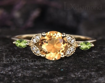 Peut inclure: Une bague en or avec une grande pierre orange ronde entourée de petits diamants. Deux pierres vertes en forme de poire sont situées de chaque côté de la pierre centrale.