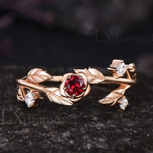 May include: Rose gold ring featuring a central red gemstone surrounded by petals. The ring has a floral design with leaves and small white gemstones. The ring is set against a dark background.