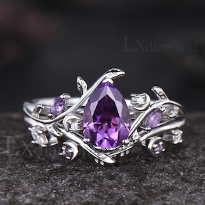 May include: A silver ring with a pear-shaped, deep purple gemstone as the focal point. The ring features a floral design with smaller purple and clear gemstones. The band has a delicate, vine-like pattern. The text "Lxdiamond" is visible.