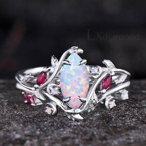 Peut inclure: Une bague en argent avec une grande pierre centrale opale ovale et irisée. La bague présente un motif floral avec des pierres d'accent rouges et blanches et le texte "Lxdiamond".