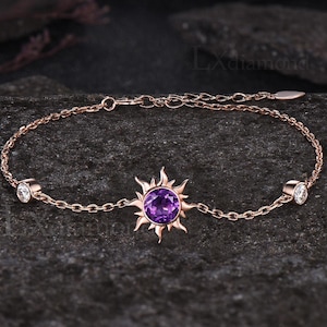 May include: Rose gold bracelet featuring a sunburst design with a central purple gemstone and two smaller white gemstones. The delicate chain bracelet is displayed on a dark, textured surface.