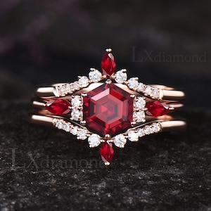 Könnte beinhalten: Ein dreiteiliger Verlobungsring aus Roségold mit einem großen roten Edelstein, der von kleineren weißen Edelsteinen umgeben ist. Der mittlere Stein ist sechseckig geschliffen.