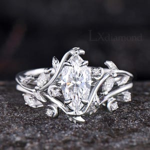 Pode incluir: Um anel de tom prateado com uma grande pedra central de corte marquise, rodeada por uma banda com design de videira metálica e pedras menores de corte marquise e redondo. O texto LXdiamond é visível no canto superior direito da imagem.