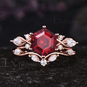 May include: A rose gold engagement ring set featuring a large, hexagonal, deep red gemstone. The ring is accented with smaller white gemstones and a delicate, leaf-like design. The set includes a matching band.
