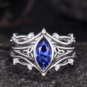 May include: A silver-toned ring featuring a central marquise-cut blue gemstone, flanked by intricate metal knotwork patterns and small clear accent stones along the band. The text LXdiamond is visible in the upper right and lower left corners.